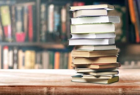 Стос книг на дерев'яній поверхні біля полиці з книгами.